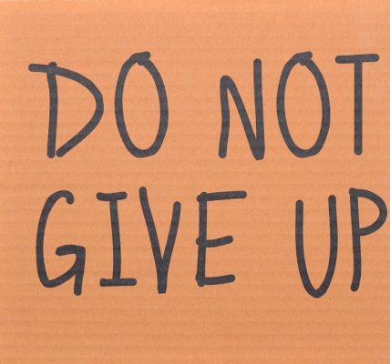 Cardboard sign reading 'Do Not Give Up' placed on green turf, symbolizing perseverance.