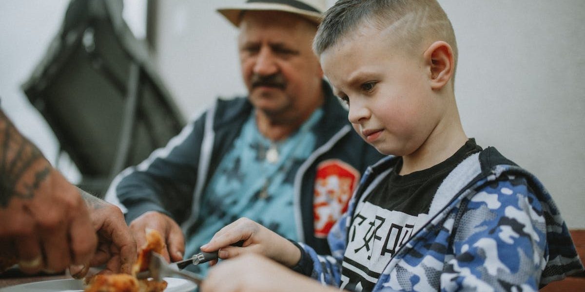 A child and his father share a meal outdoors, enjoying grilled food together.