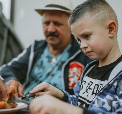 A child and his father share a meal outdoors, enjoying grilled food together.